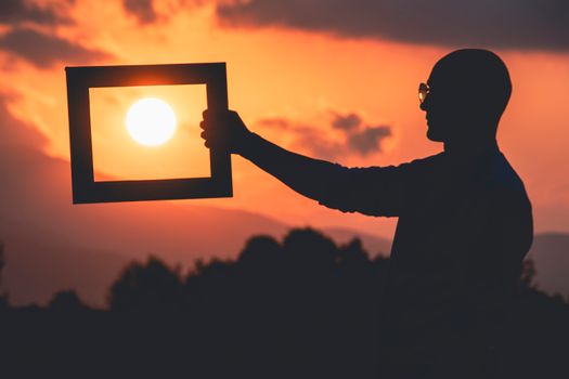 Silhouette of a man holding a frame enclosing the sun at sunset, creating a unique composition.