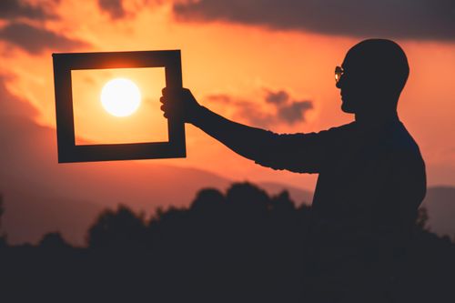 Free Silhouette of a man holding a frame enclosing the sun at sunset, creating a unique composition. Stock Photo