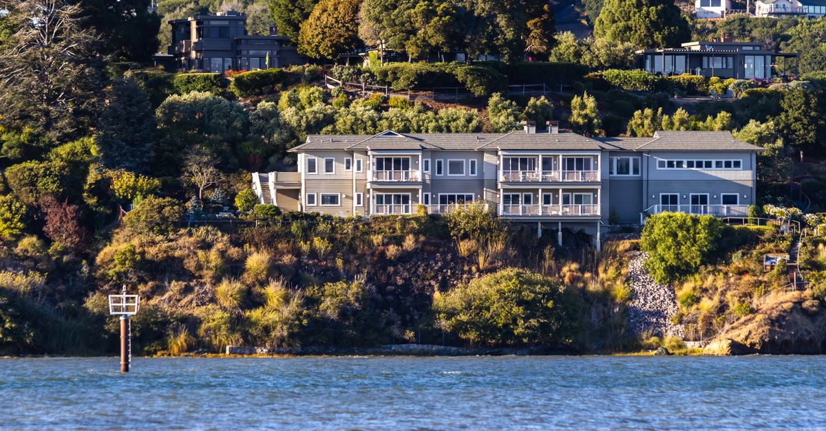 A scenic view of a large waterfront mansion surrounded by trees and greenery, overlooking the sea.