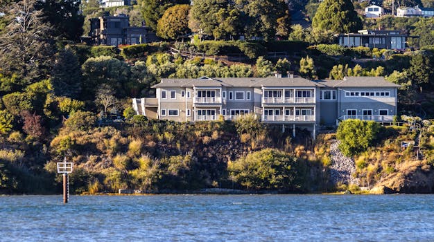 A scenic view of a large waterfront mansion surrounded by trees and greenery, overlooking the sea.