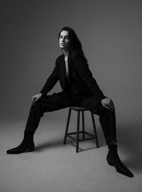 Stylish portrait of a woman in a suit and ankle boots sitting on a stool in black and white.