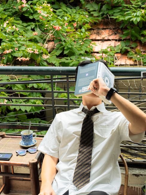 Young man with book on his face, relaxing on a balcony surrounded by greenery.