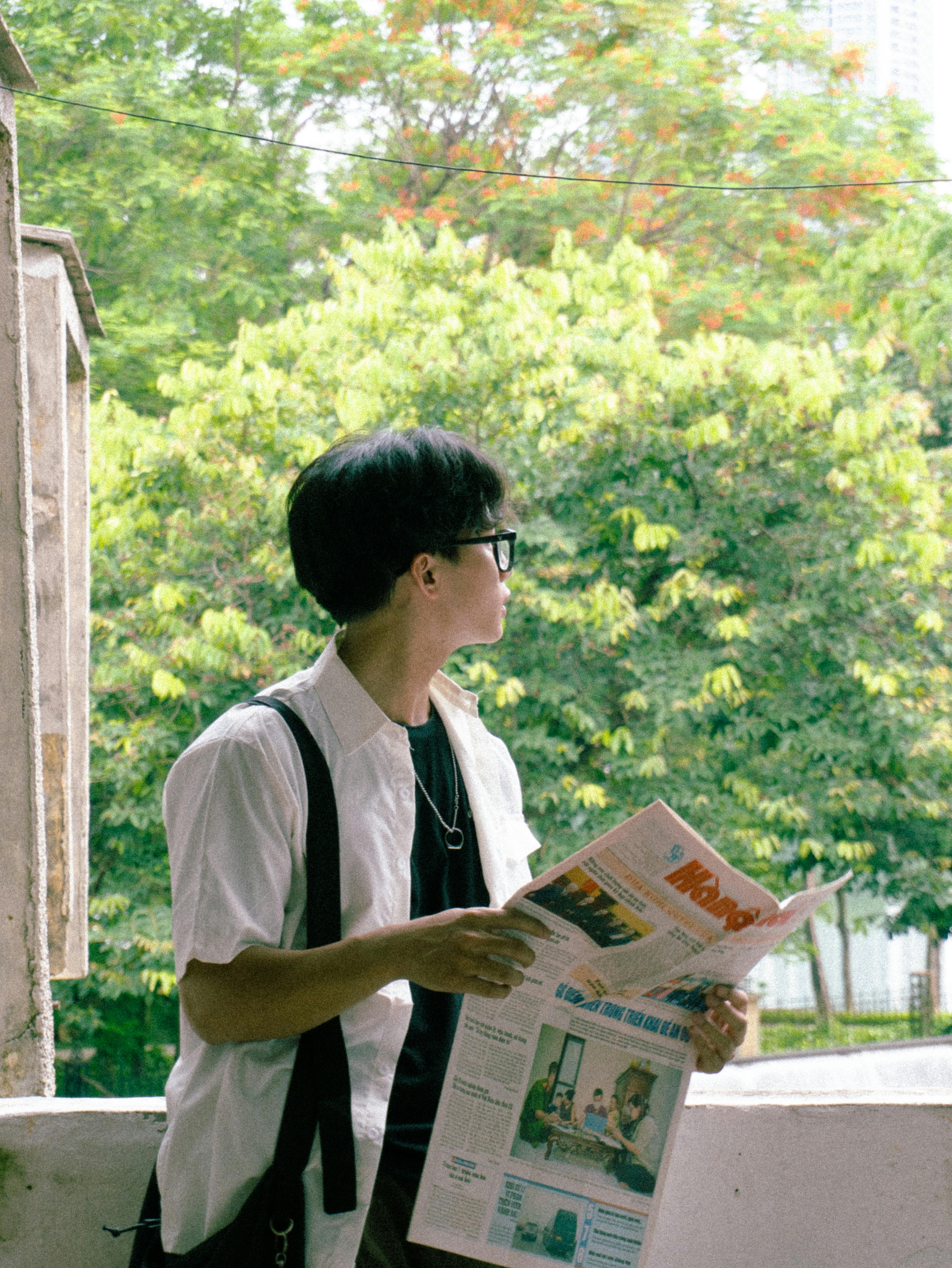 Young man with eyeglasses reading newspaper on balcony with lush trees in the background.