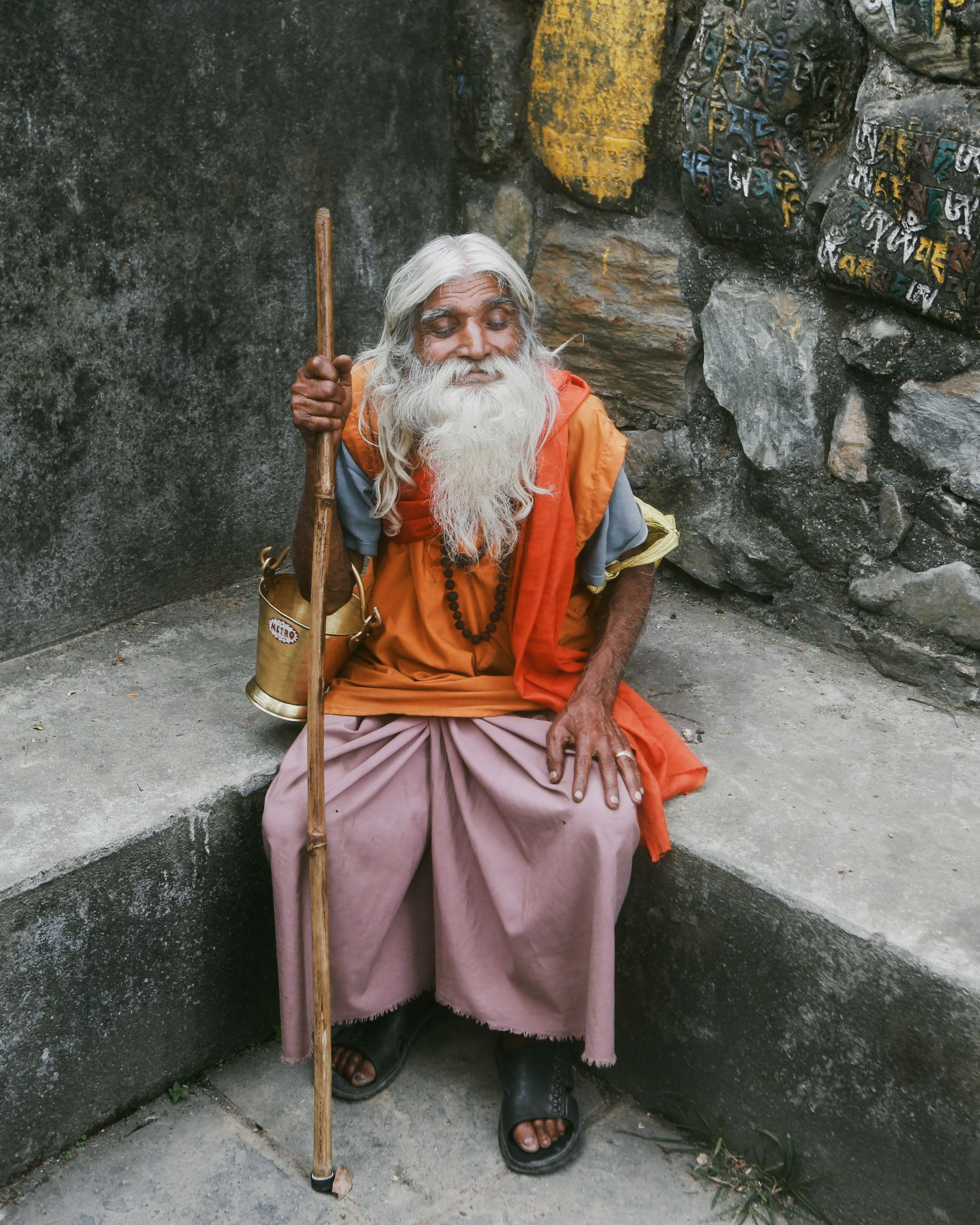 An elderly man, dressed traditionally, sits on a bench holding a wooden staff.