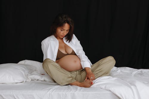 Pregnant Woman in White Shirt and Beige Pants Sitting on a Bed