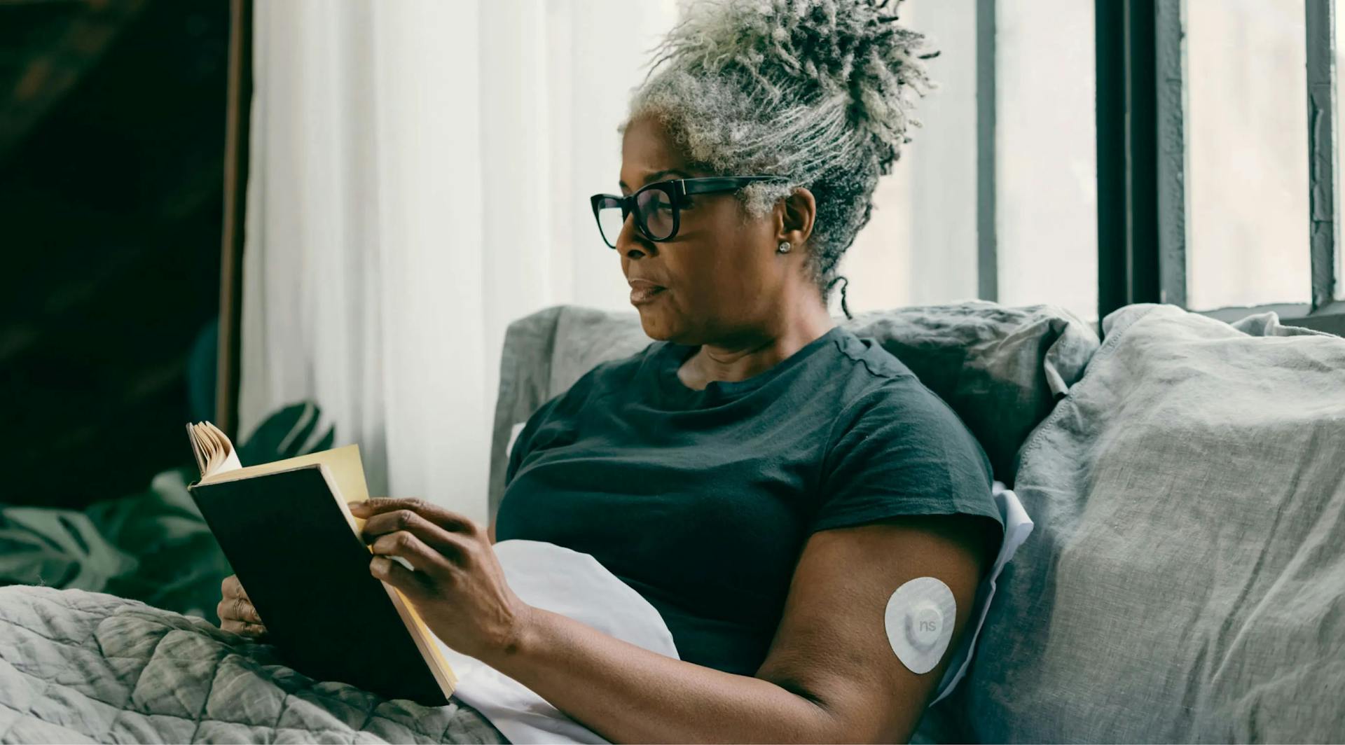 Senior woman with gray hair reading a book indoors wearing a glucose monitoring sensor.