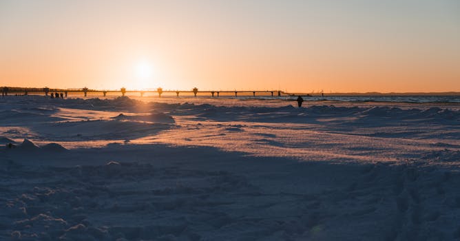 Serene winter sunset reflecting over a snowy beach with distant pier view.