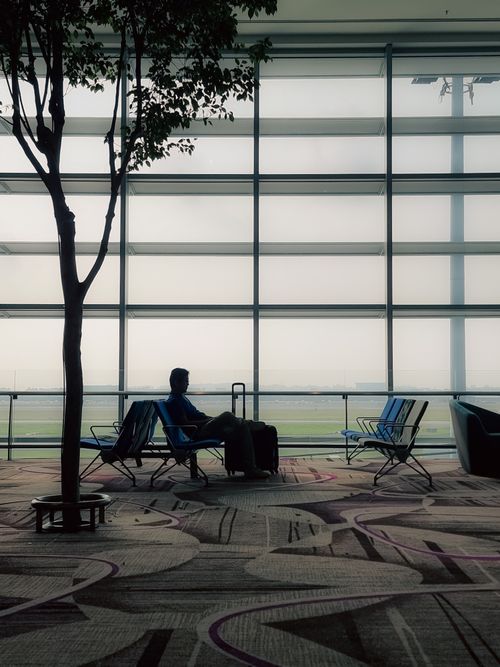 Free Silhouetted traveler sitting in airport lounge with luggage, near large windows. Stock Photo
