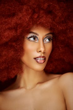 Vibrant portrait of a woman with artistic makeup and a bold red afro wig in a studio setting.