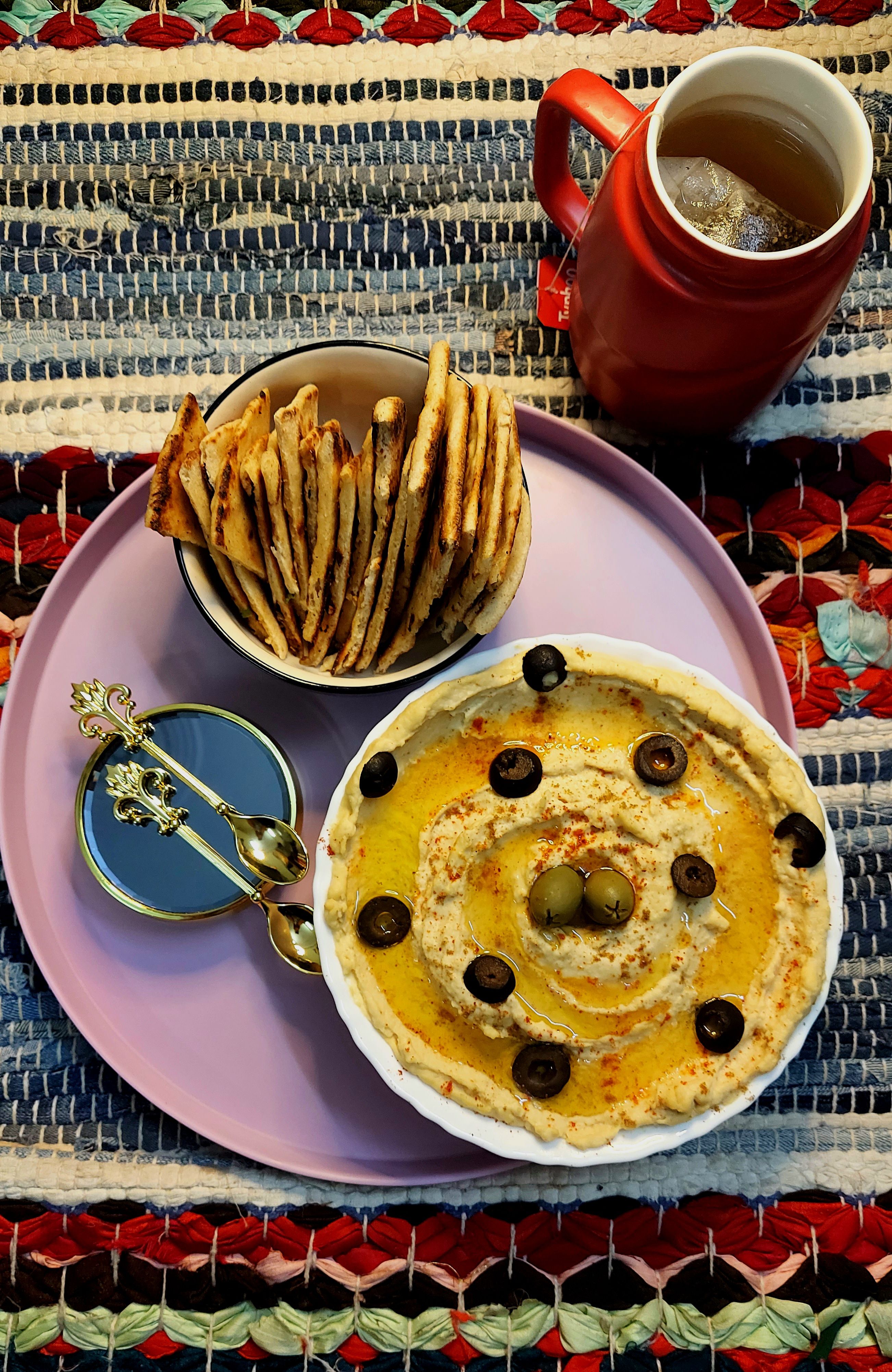 Top view of a hummus platter with olives, pita bread, and tea on a woven mat.
