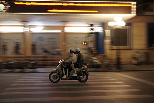 Blurred motion of people riding a motorbike across a Hanoi city street at night.