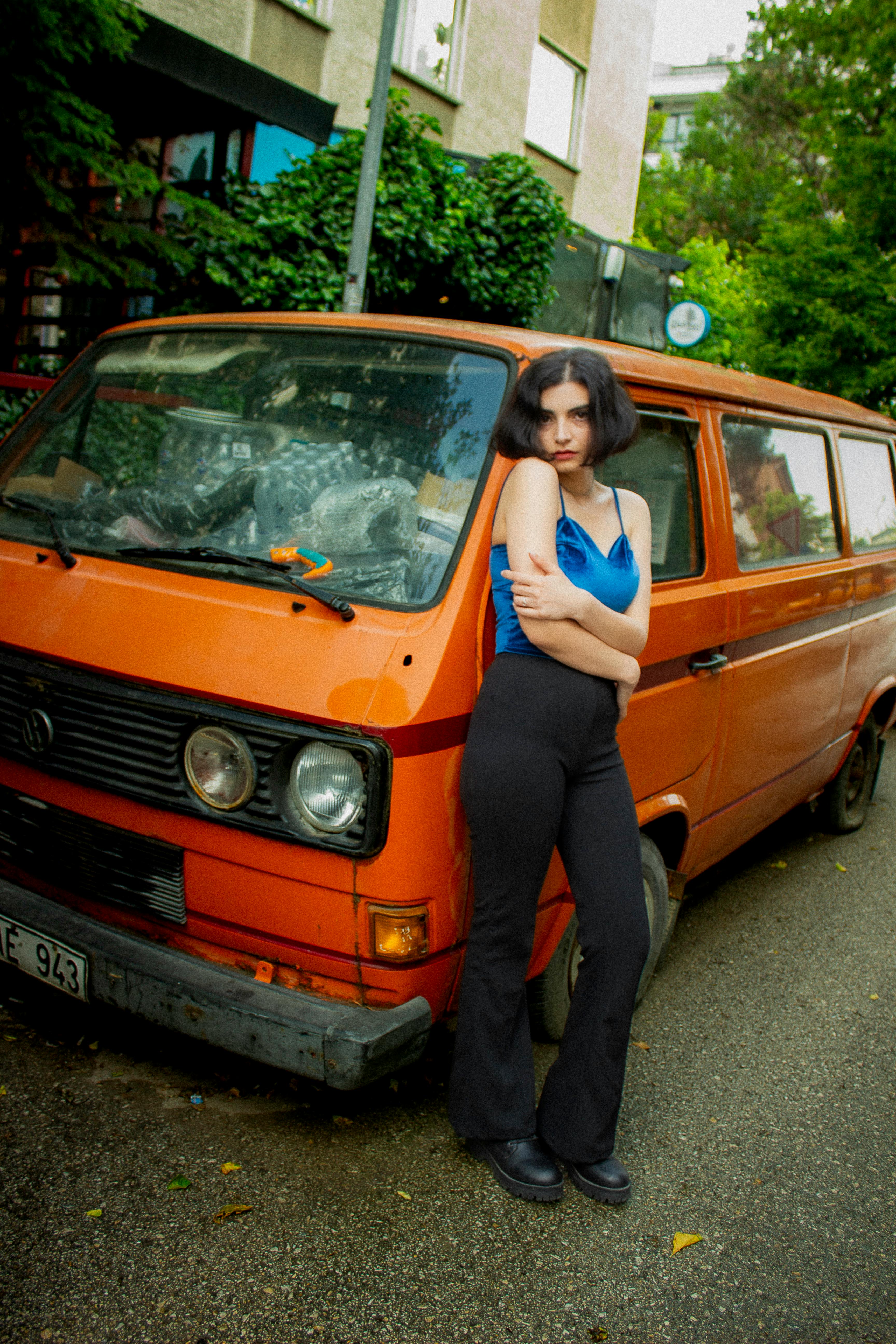 A stylish young woman stands confidently by a vintage orange van in an urban setting.