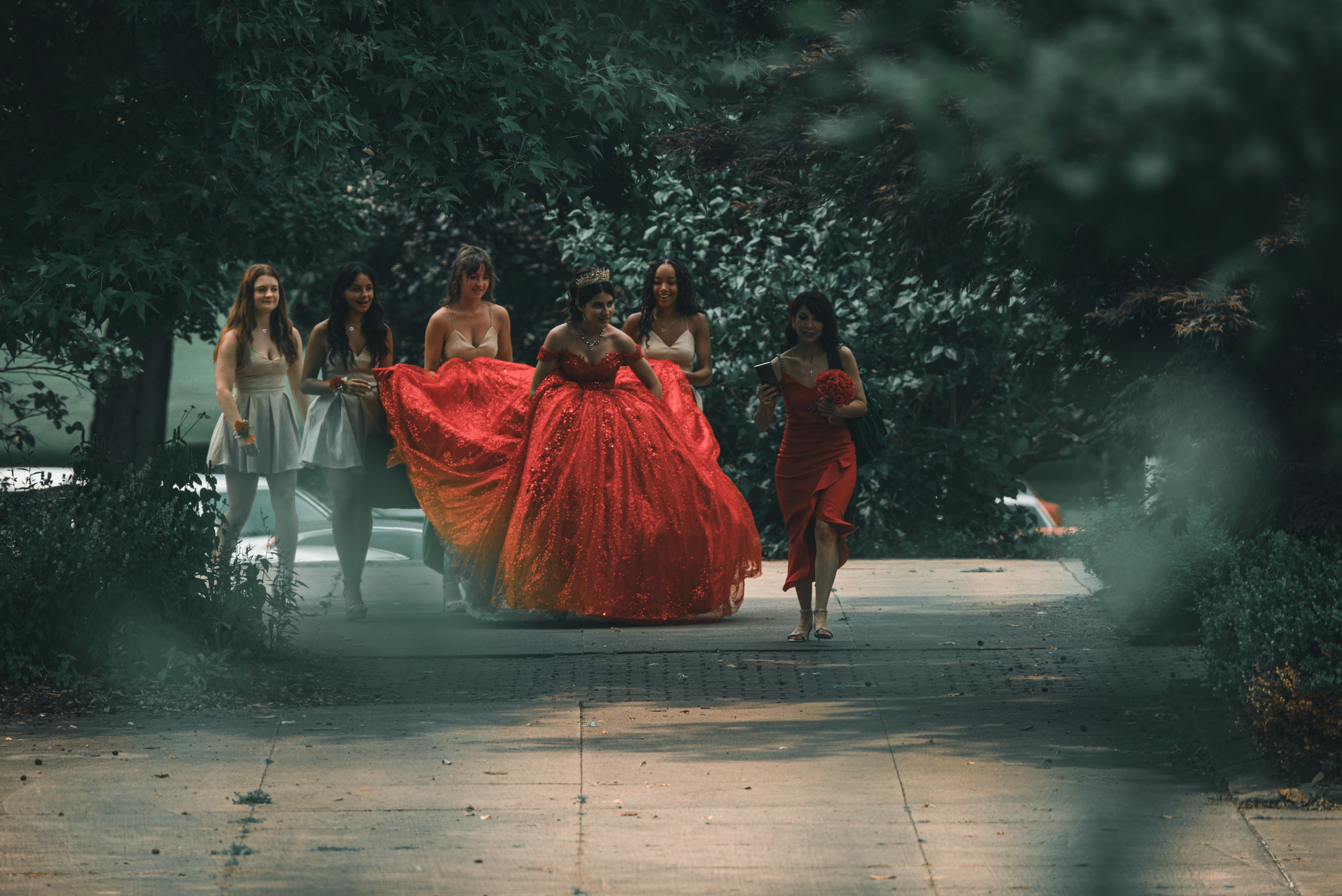 Bride in stunning red wedding gown with bridesmaids walking through lush outdoor setting.