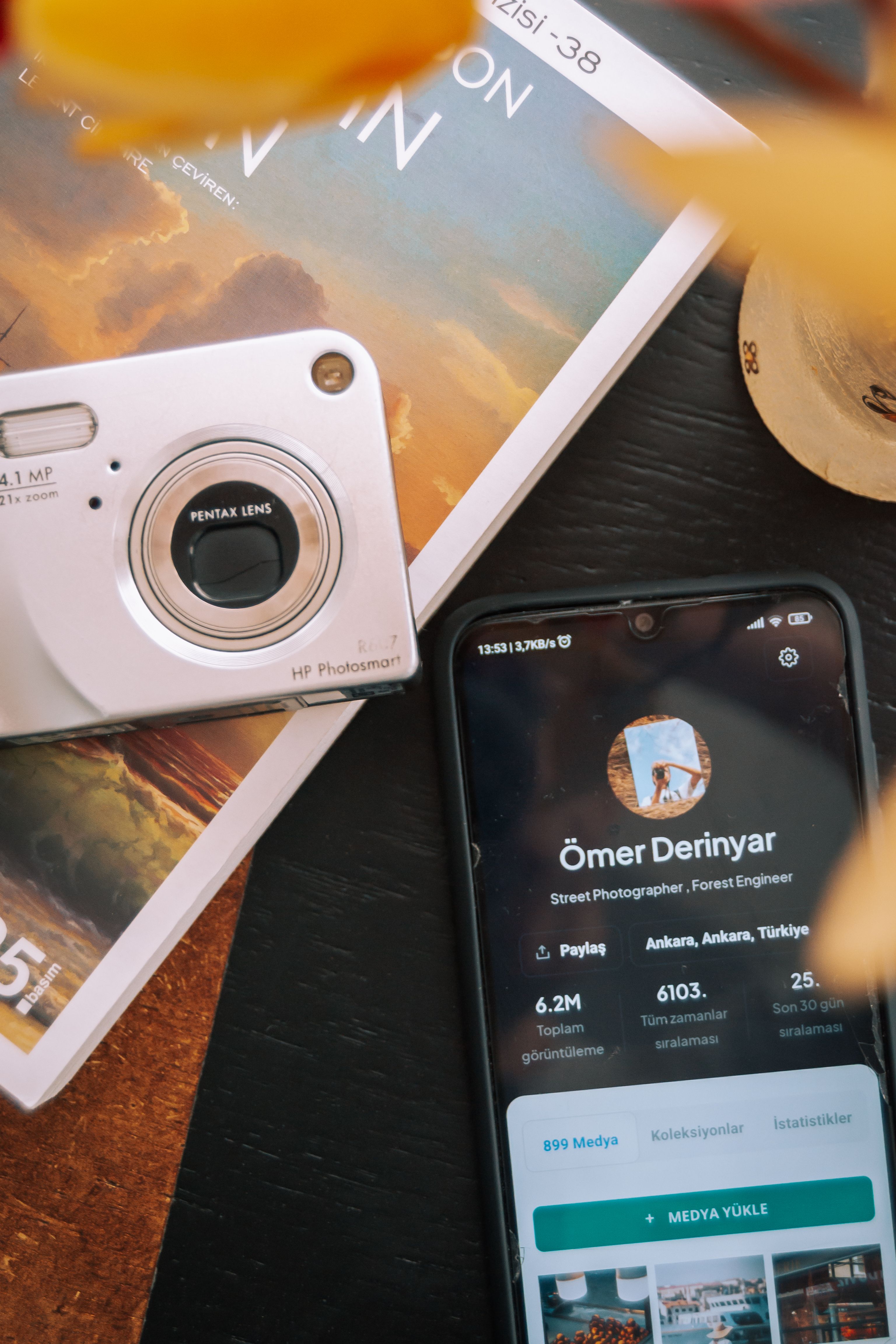 A close-up of an old digital camera next to a smartphone displaying a profile page on a wooden table.