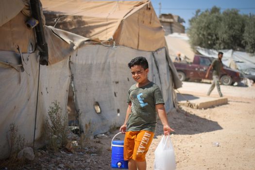 A young boy carries water at a Syrian refugee camp under a sunny sky.