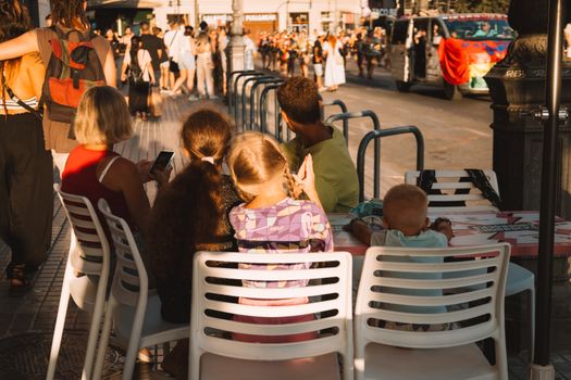 A family seated at a cafe watches a lively parade pass by in the city streets.