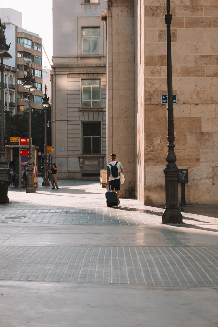 Man Walking With Suitcase And Backpack Near Building Wall