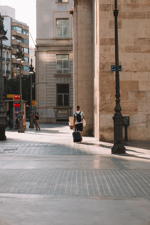 Free A man with a suitcase walks down a sunlit urban street, showcasing city life. Stock Photo
