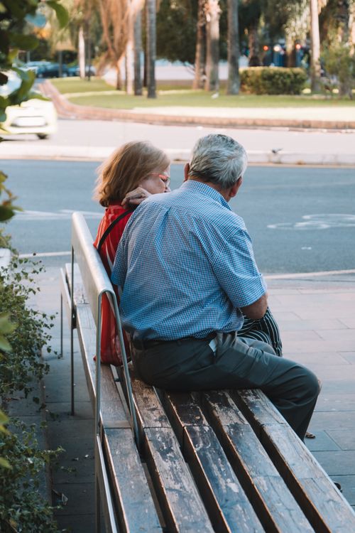 Free Elderly man and woman sitting on a bench in a sunny urban setting, enjoying a relaxing moment. Stock Photo