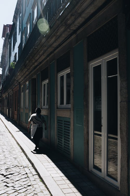 Person Walking near Building in Shadow by Sunlit Street