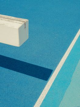 A minimalist view of a blue running track with white lines and a wooden beam casting a shadow.