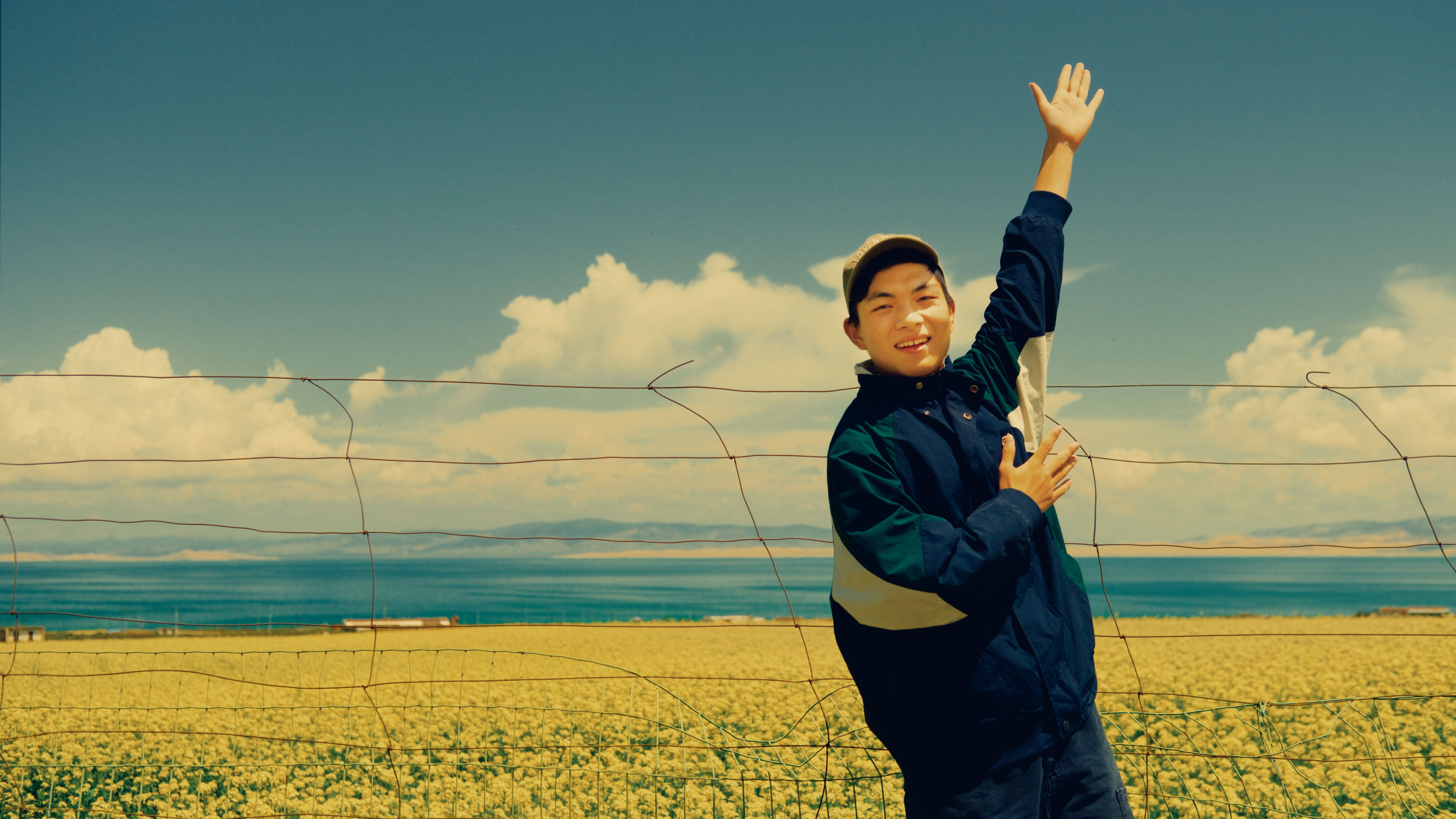 Smiling young man posing near a rapeseed field with vibrant blue sky and scenic lake view.