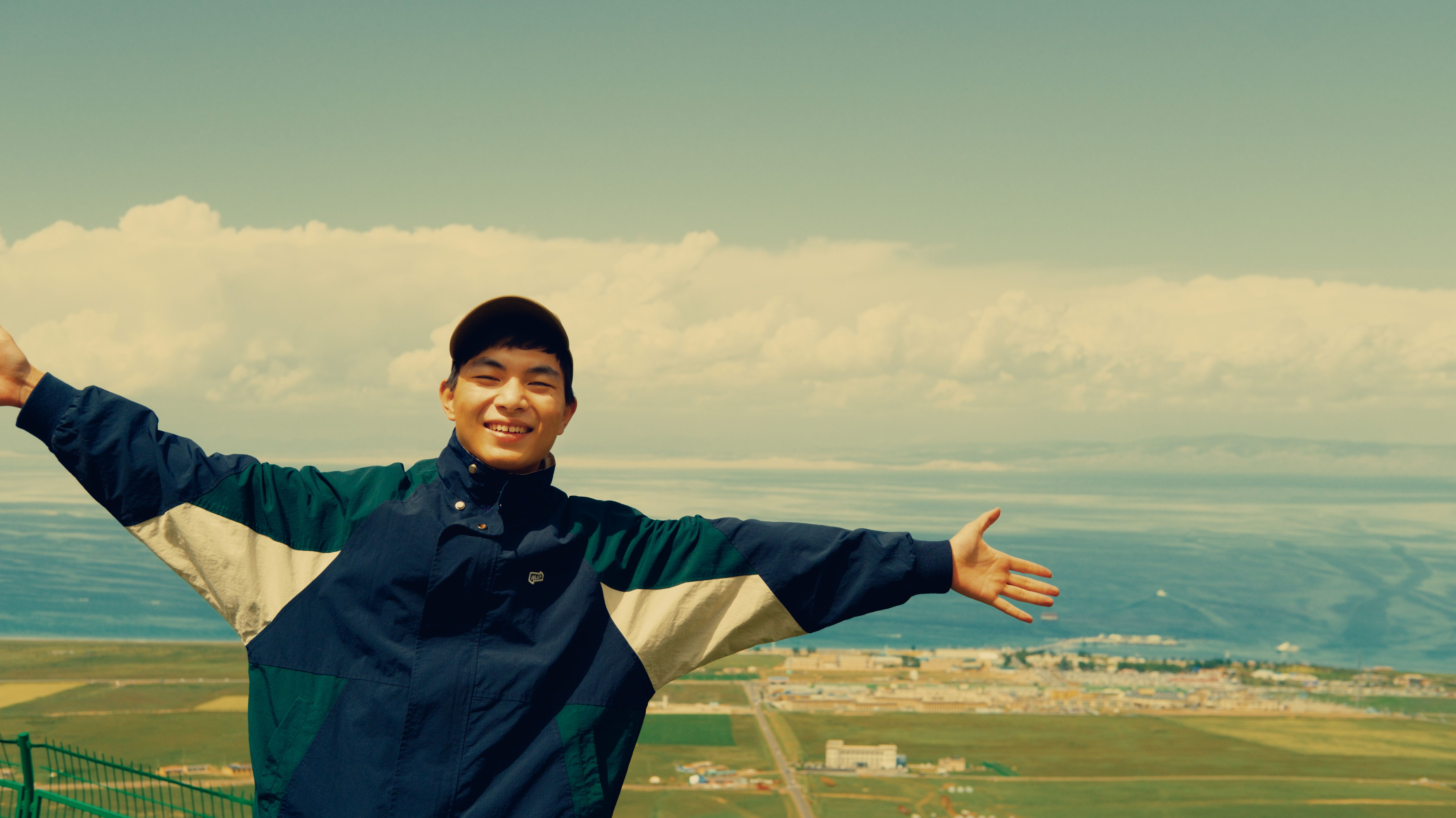 A young man stands with arms spread, smiling in a scenic outdoor setting.