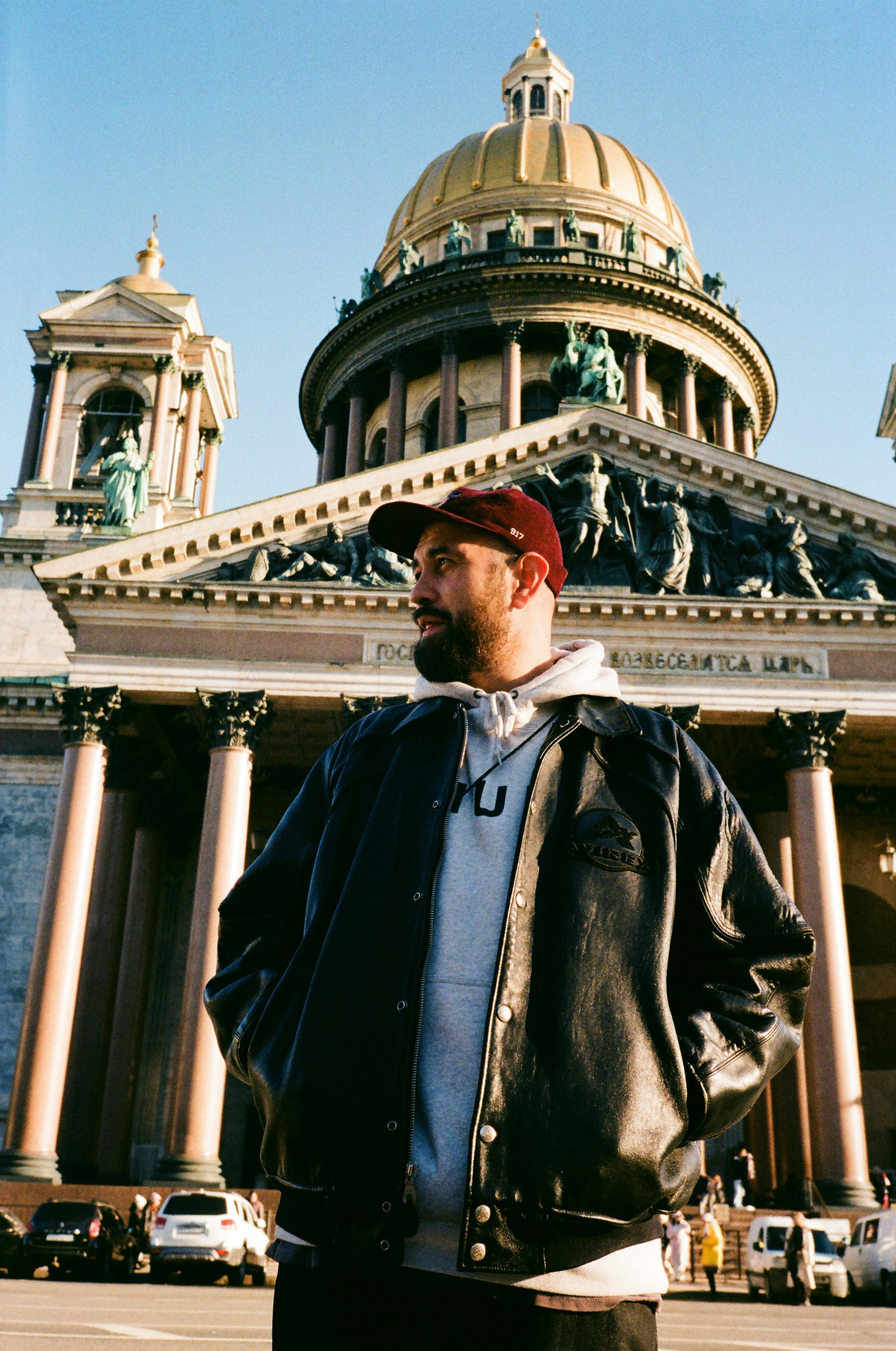 A man in a black jacket stands before a historic cathedral with a golden dome in the city.