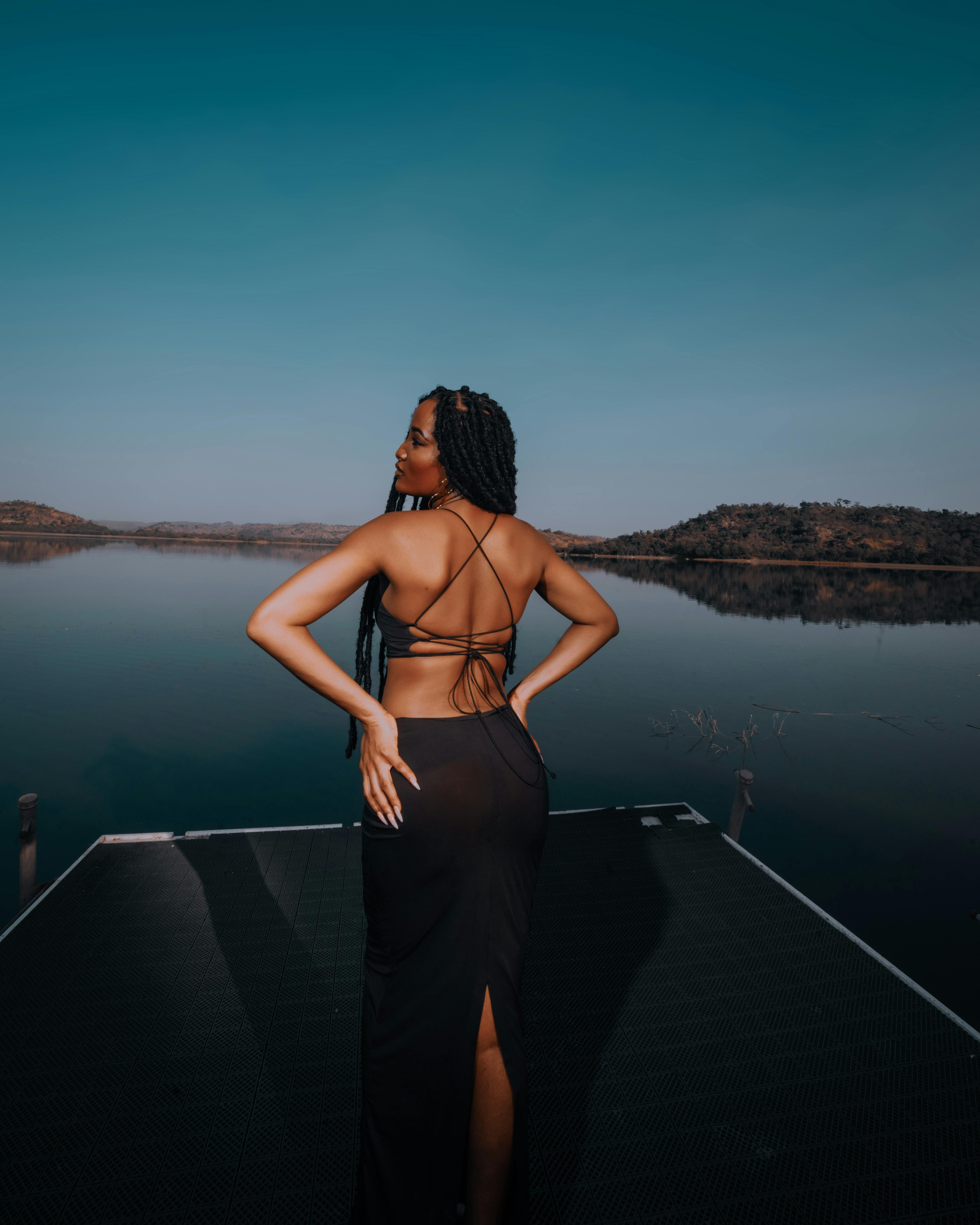 A stylish woman in a black dress stands gracefully on a pier overlooking a tranquil lake.