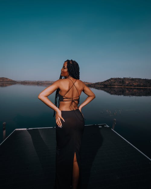 Woman in Black Dress Standing on Pier on Lake