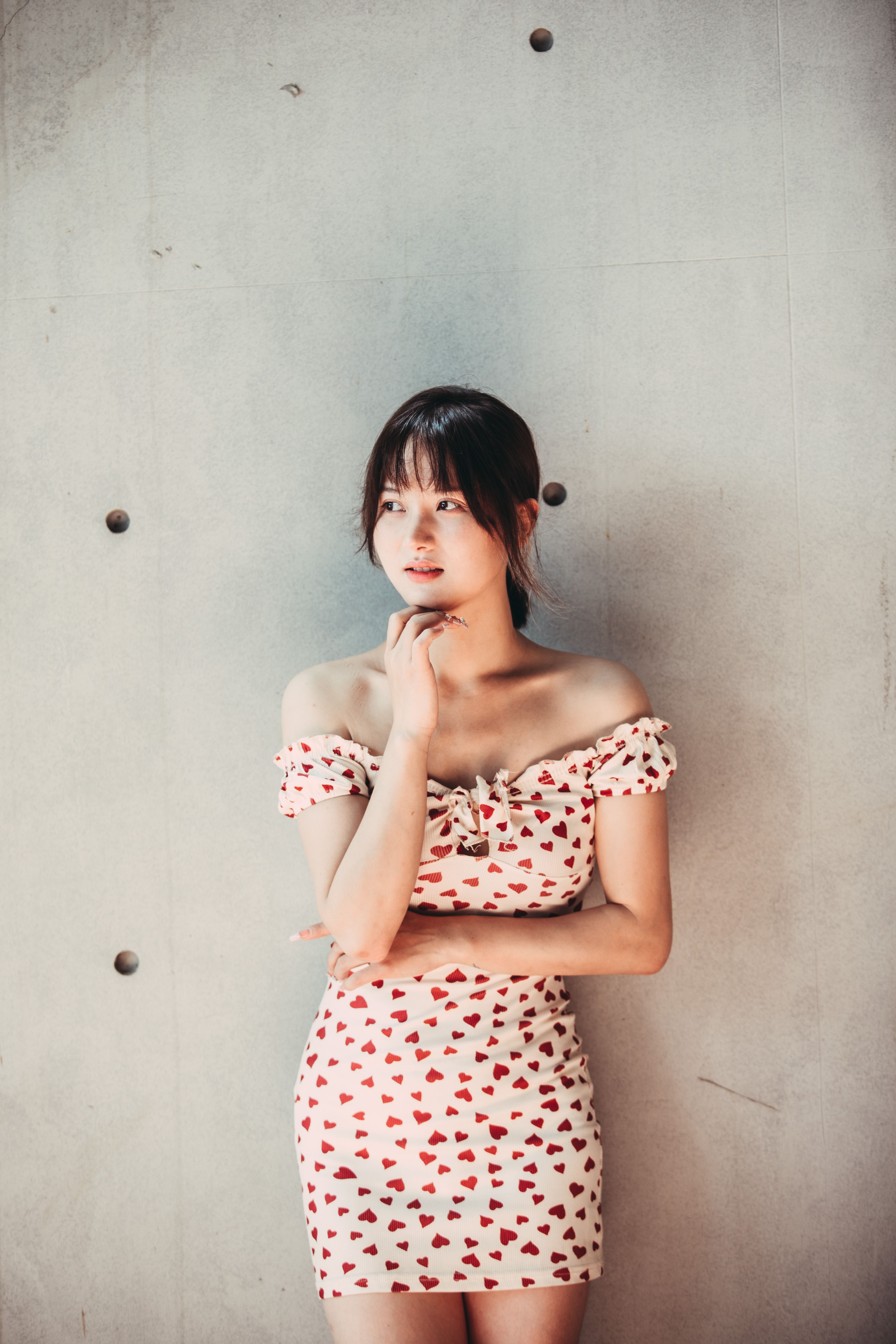 A young woman in a heart-patterned dress poses elegantly against a neutral wall, showcasing summer fashion style.