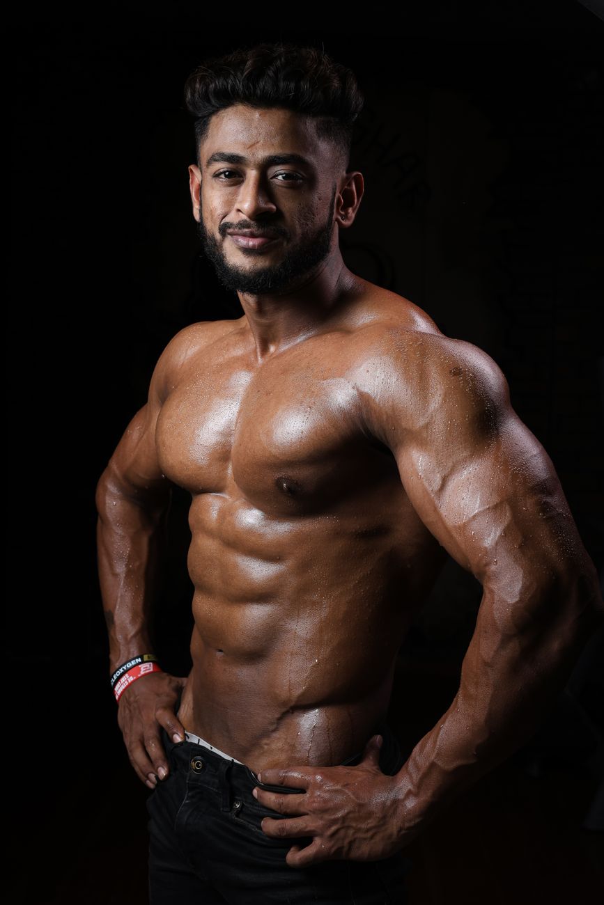 A muscular shirtless man with a beard confidently poses in a studio, showcasing defined muscles and a wristband.
