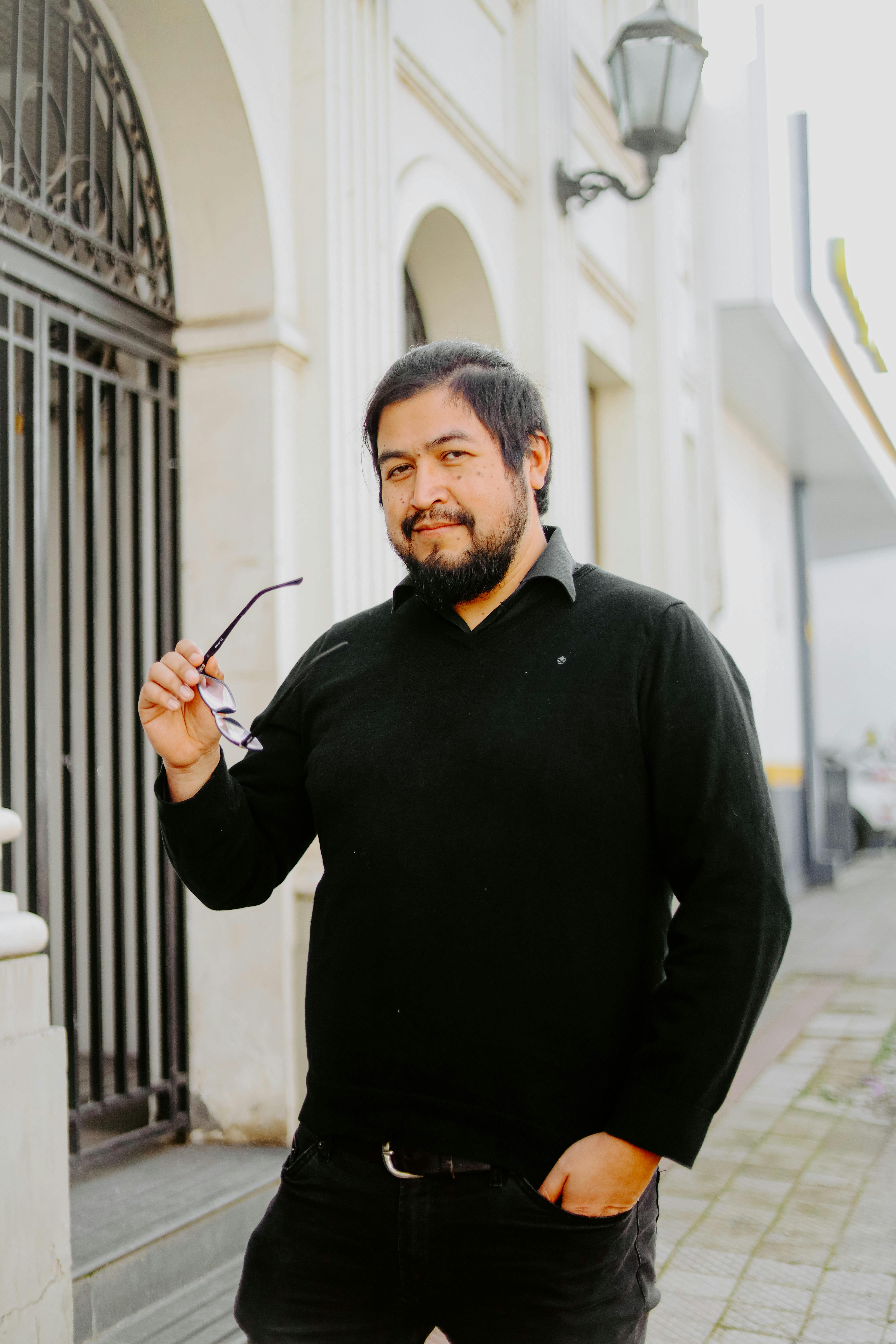 Casual portrait of a bearded man standing near an urban metal gate holding eyeglasses.