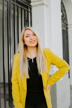 Portrait of a confident businesswoman with long blonde hair wearing a stylish yellow blazer, smiling outdoors.