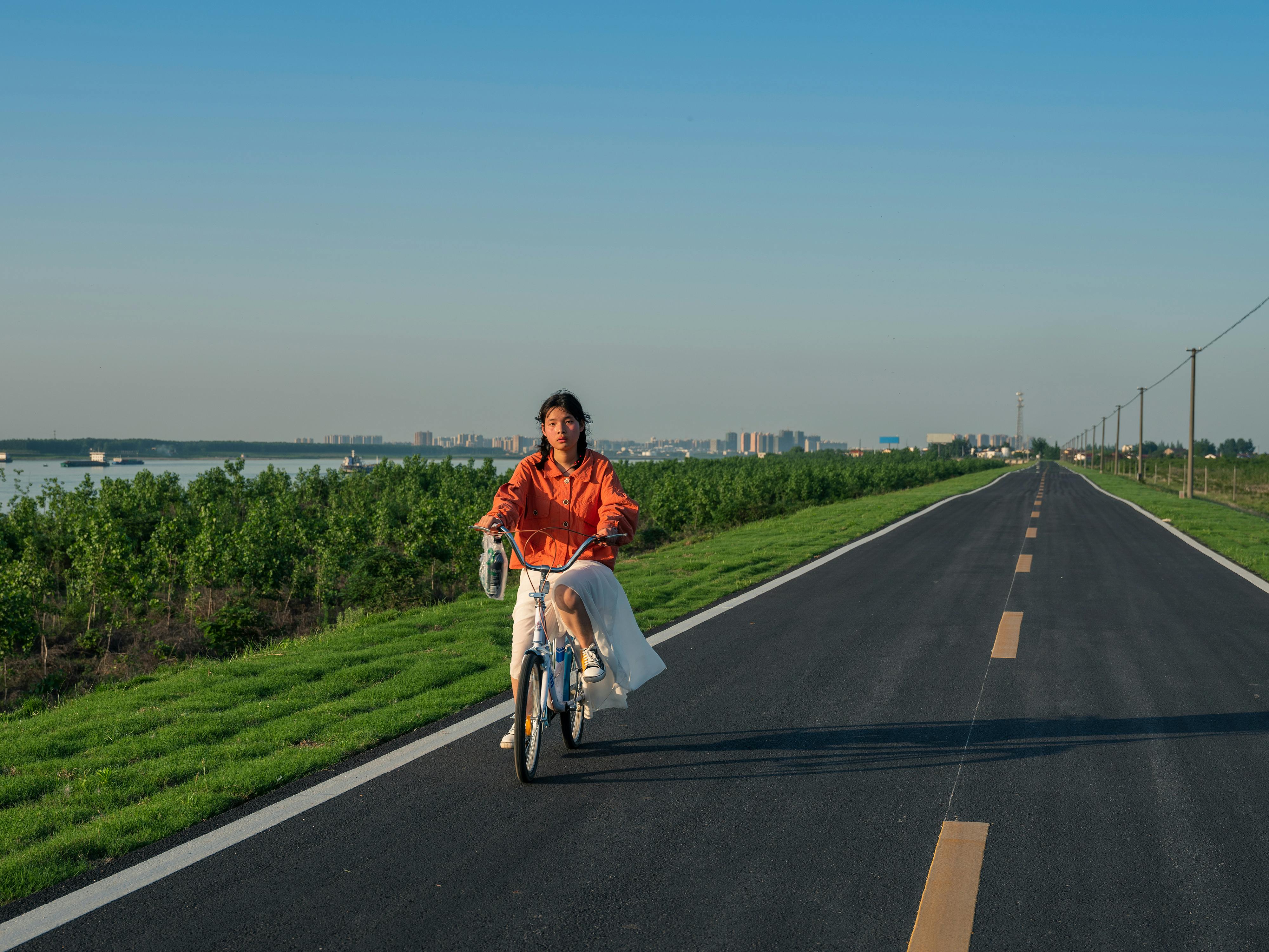 A woman rides a bicycle on a straight rural road under clear blue sky.