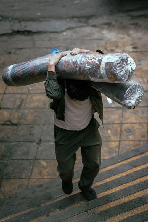 Free A man carries heavy rolled carpets on his back up concrete steps in an urban environment. Stock Photo