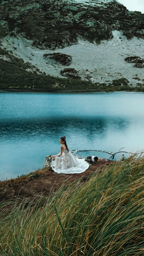 Woman in a White Dress Sitting by the Body of Water in the Valley 