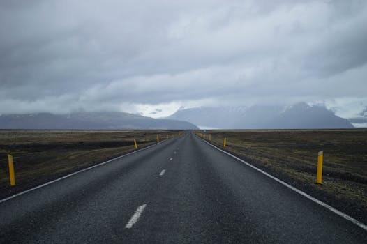 A long empty road stretches towards snow-capped mountains under a cloudy sky in Iceland.
