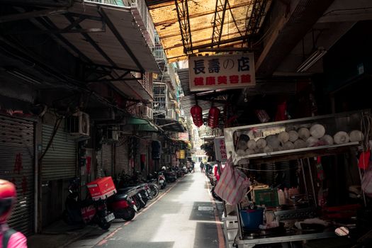 A vibrant urban alley featuring parked motor scooters and numerous shops with signage, evoking a lively street market vibe.