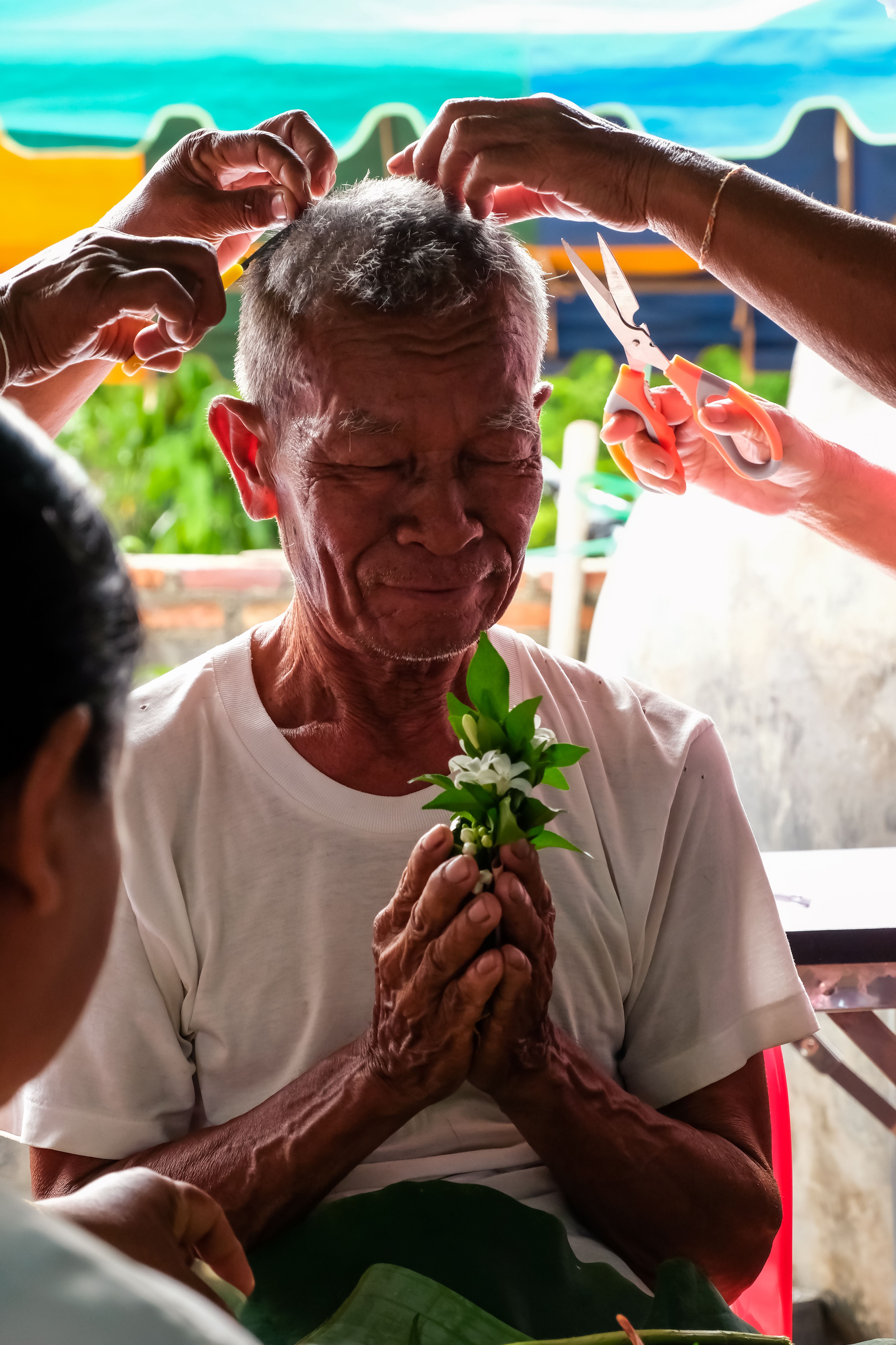 An elderly man holds flowers with eyes closed during an outdoor haircut ceremony. - Fotografer Sidoarjo