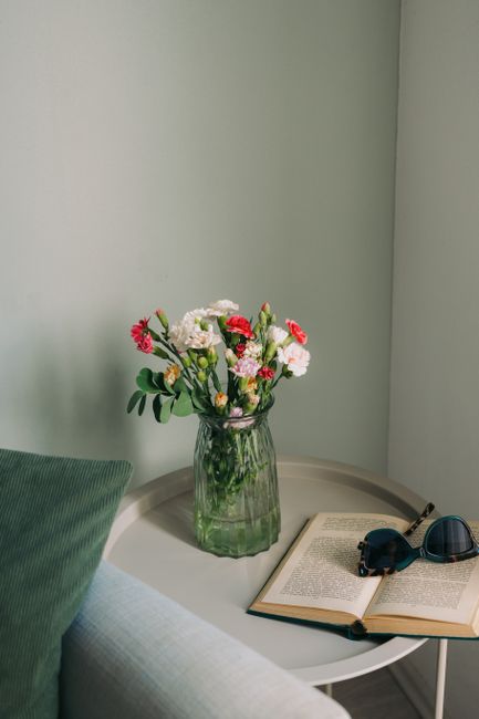 A vase of colorful carnations beside an open book and sunglasses on a table.