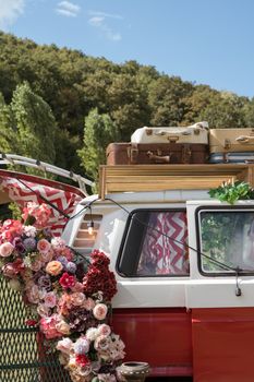 Vintage red van adorned with colorful flowers and luggage, set against nature.