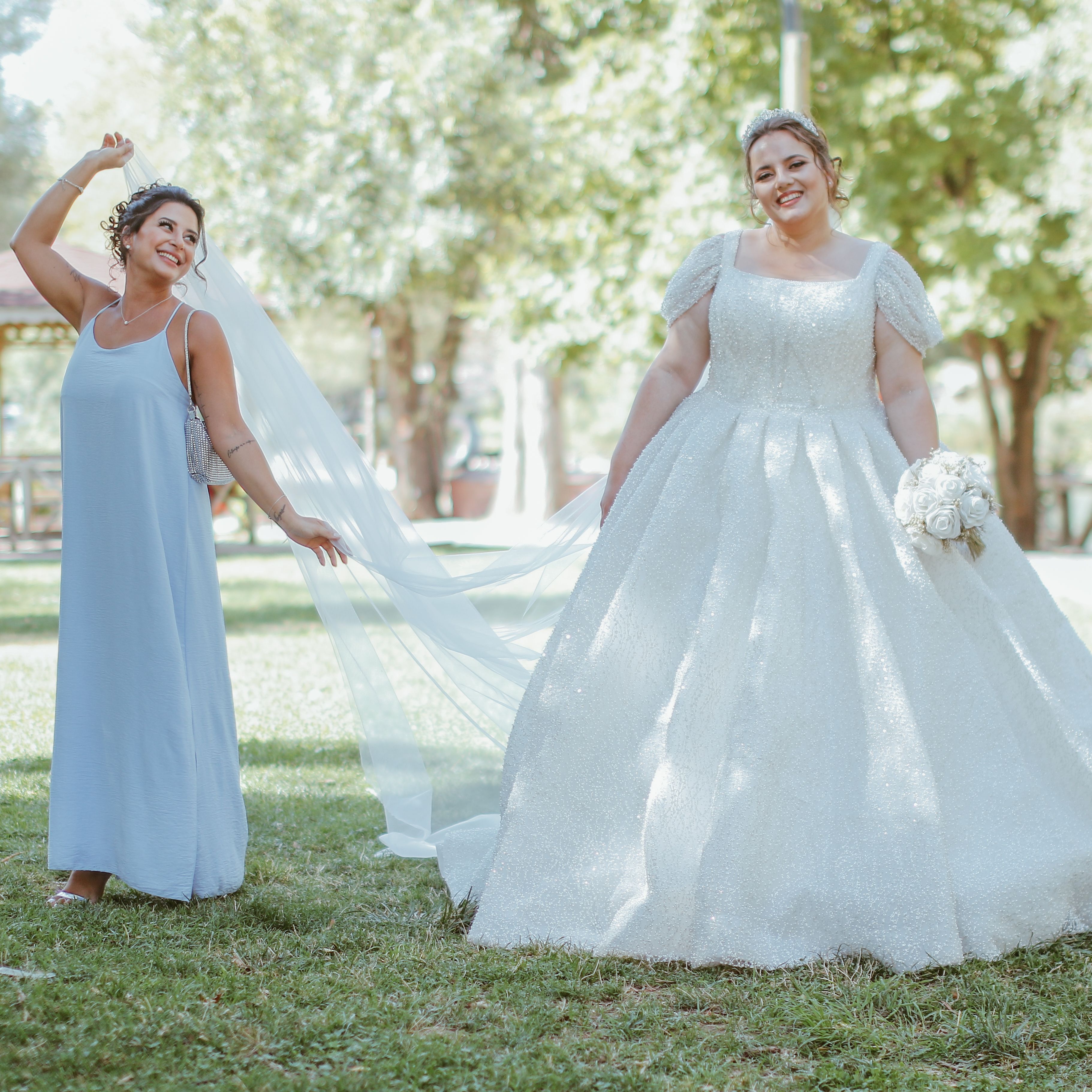 A bride and bridesmaid enjoying a sunny day in the park, showcasing a joyful wedding moment.