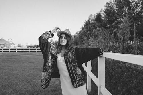 Woman in Cowboy Hat and Ethnic Jacket Posing near Fence in Field
