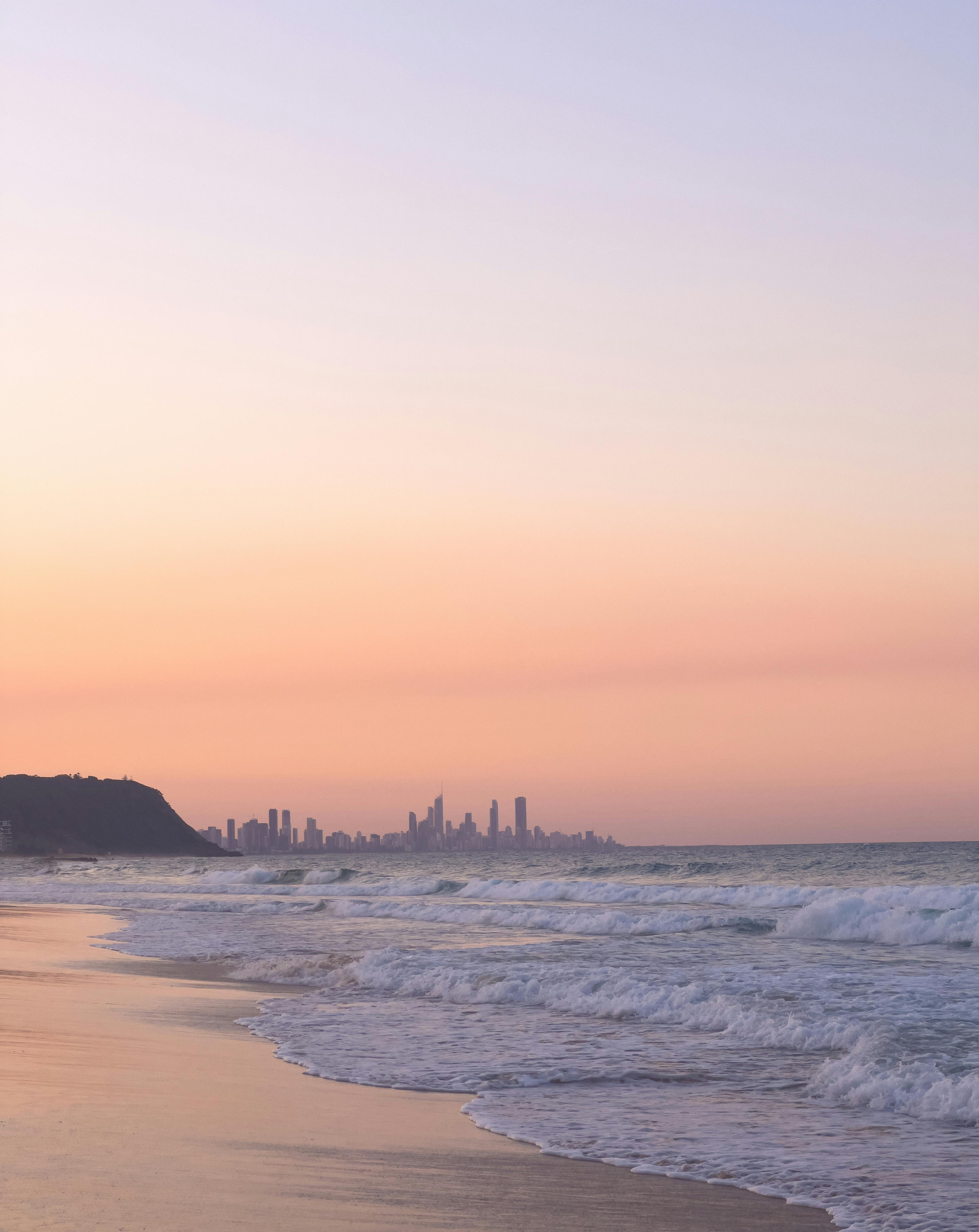 Serene view of Gold Coast beach at sunset with city skyline in the distance and waves gently lapping the shore.