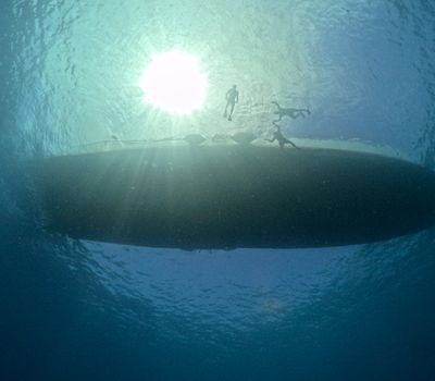 Silhouetted swimmers explore underwater near a boat under sunlight.