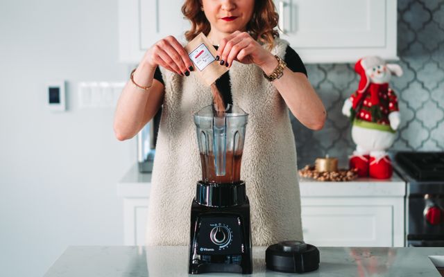 Woman pouring a sachet into a tall Vitamix blender jug on a black base with a dial, in a bright white kitchen