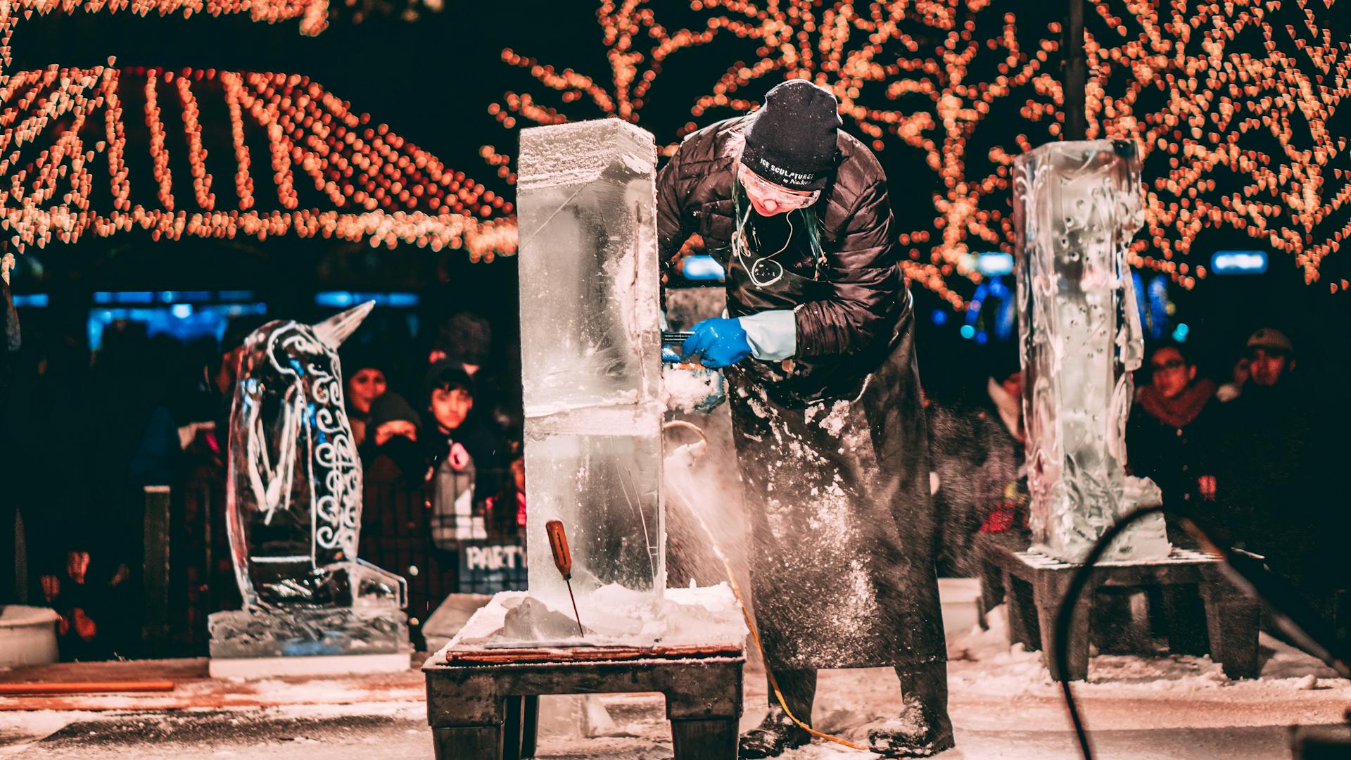 An artist carves an ice sculpture at a lively winter festival under bright lights.