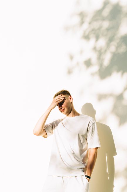 A young man in a white t-shirt relaxes outdoors, shielding his eyes from sunlight.