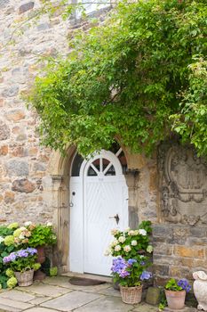 White door of a rustic stone cottage adorned with vibrant flowers and lush greenery.